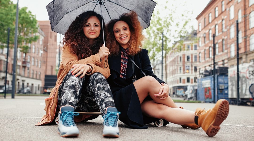 two women sitting in the street with a black umbrella posing on a skateboard